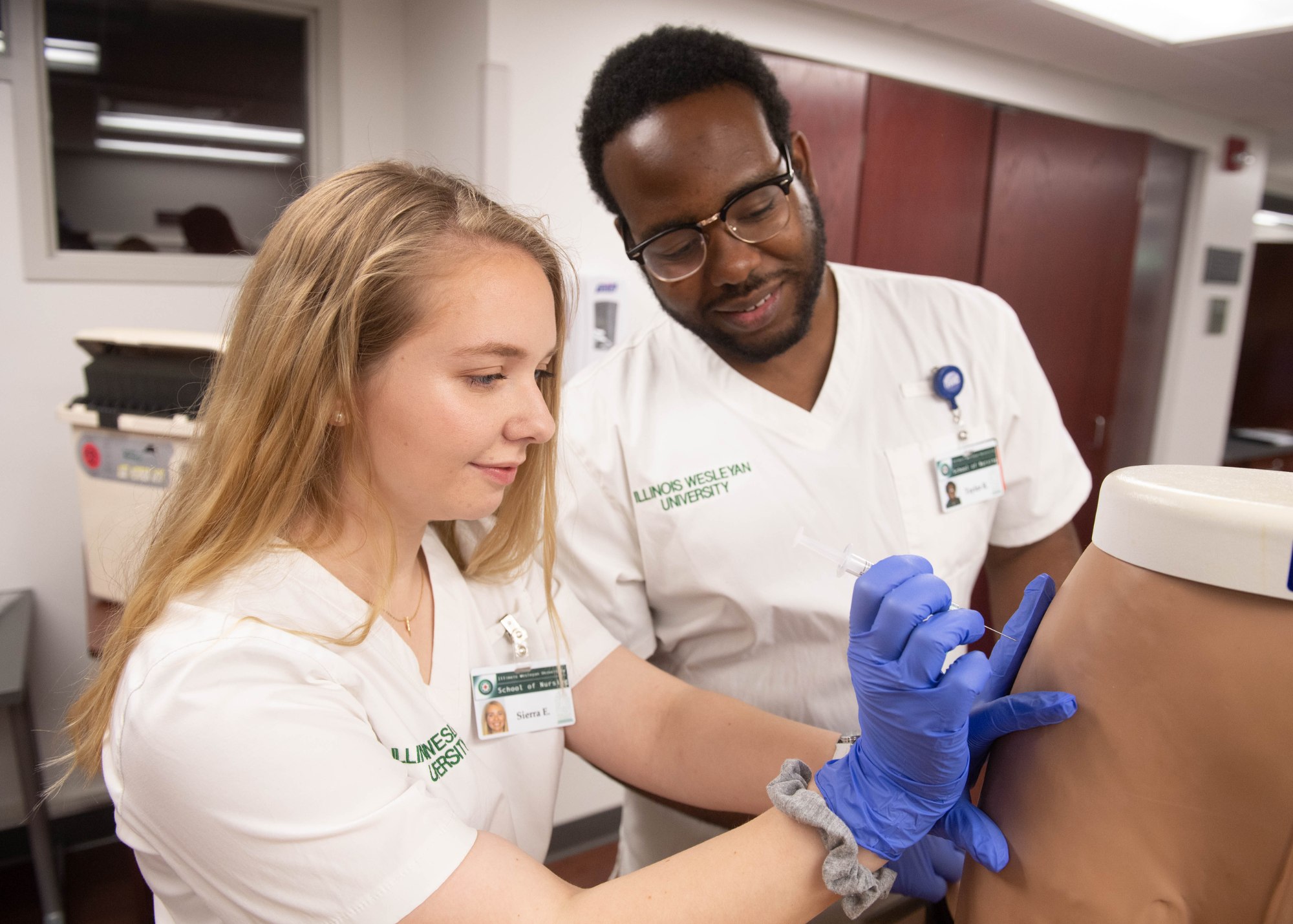 Two nursing students in scrubs practice injecting patient on a training model at Illinois Wesleyan University