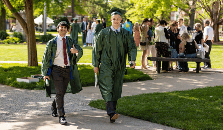 students in cap and gown walking across IWU quad after graduation, one giving thumbs up