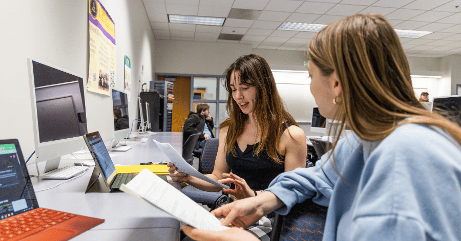 Two students speak during class holding papers and facing laptops
